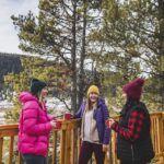 Three friends drink warm drinks on the patio overlooking backcountry scenery at Mount Engadine Lodge